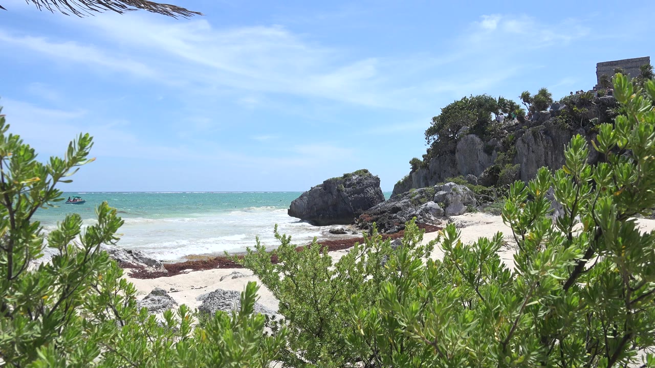 Opening frame of Tulum, Mexico coastline with green shrubs in the foreground, pale beach and turquoise surf behind them, a rocky cliff, a small stone structure above it, and no visible people.
