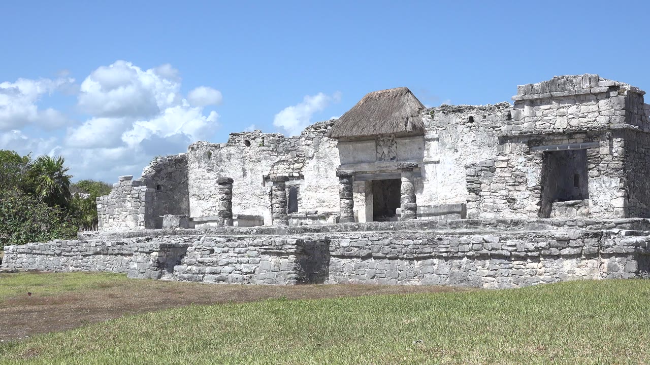 At 0:00, a weathered stone building in Tulum, Mexico stands behind grass and low stone steps under a blue sky, with no visible people.