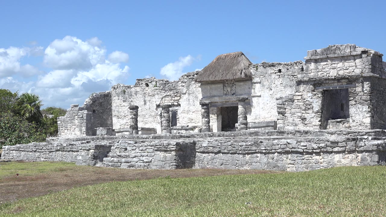 Wide view of a pale stone ruin in Tulum, Mexico with steps, wall openings, grass foreground, blue sky, clouds, and no visible people at the start of the clip.