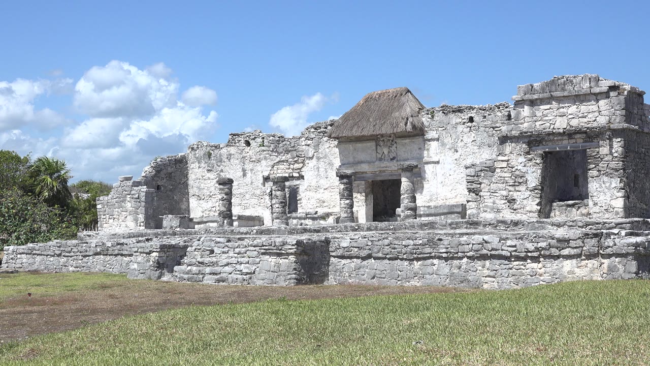 Second frame of the Tulum, Mexico ruin showing the central doorway, broken stone walls, right-side window opening, grass foreground, and no visible people.