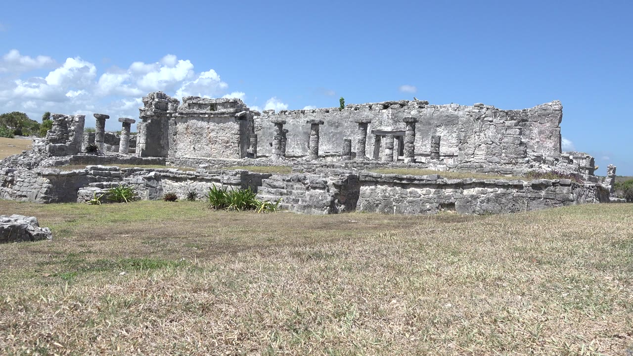 Opening frame of Tulum, Mexico stone ruins behind a dry lawn with scattered foreground rocks, pale masonry, white clouds, and no visible people.
