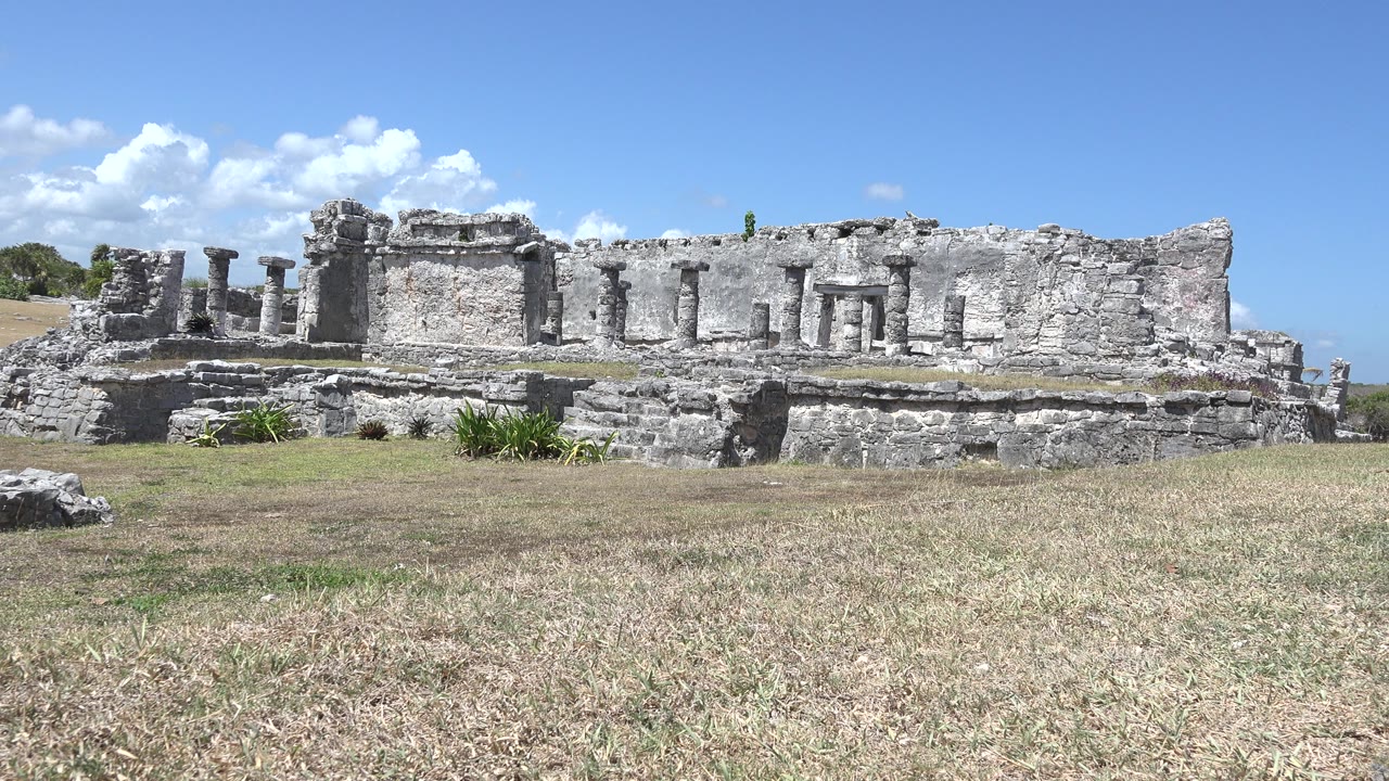 Tulum, Mexico stone ruins stretch across the frame behind dry grass and scattered rocks, with no visible people at the start of the clip.