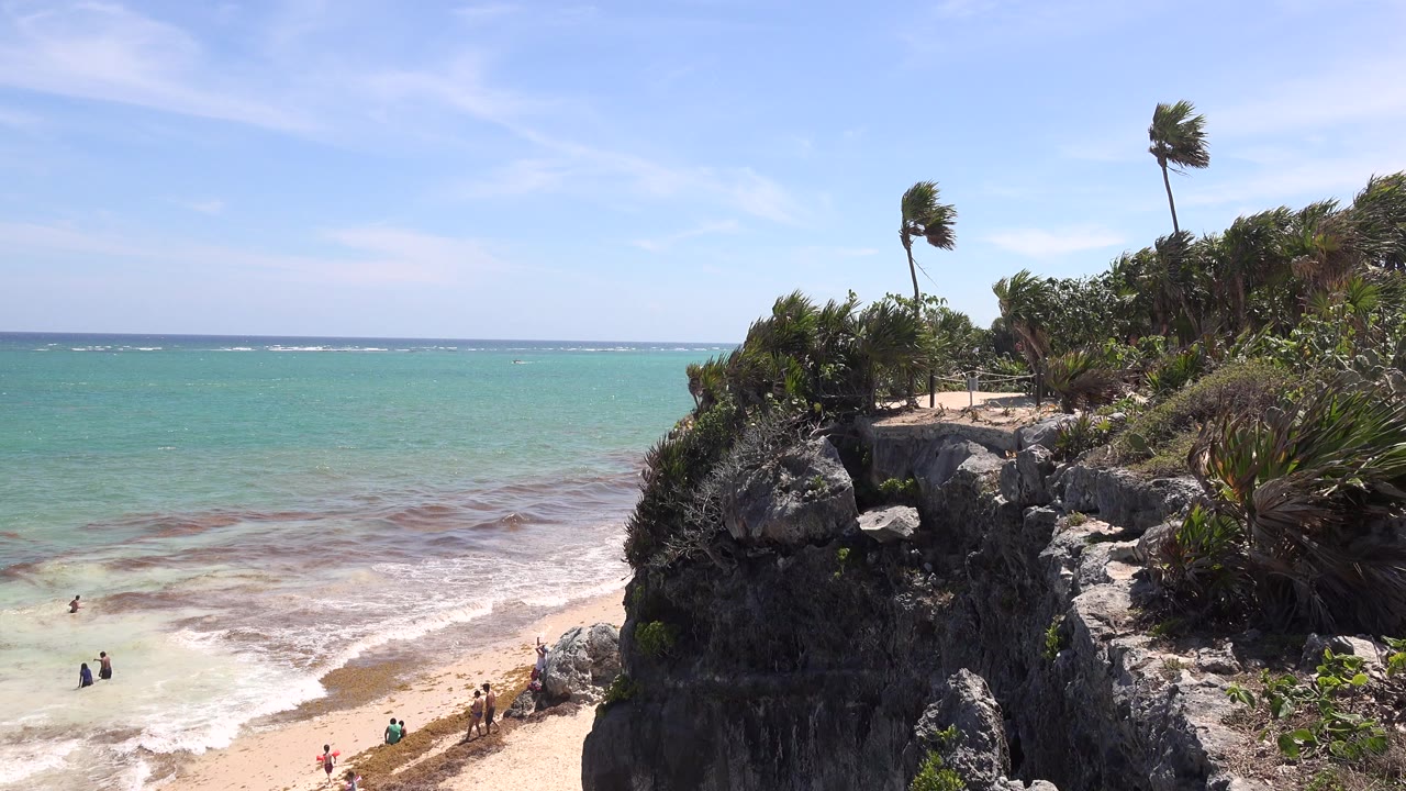 Rocky bluff above turquoise beach water with palm trees and distant beachgoers.