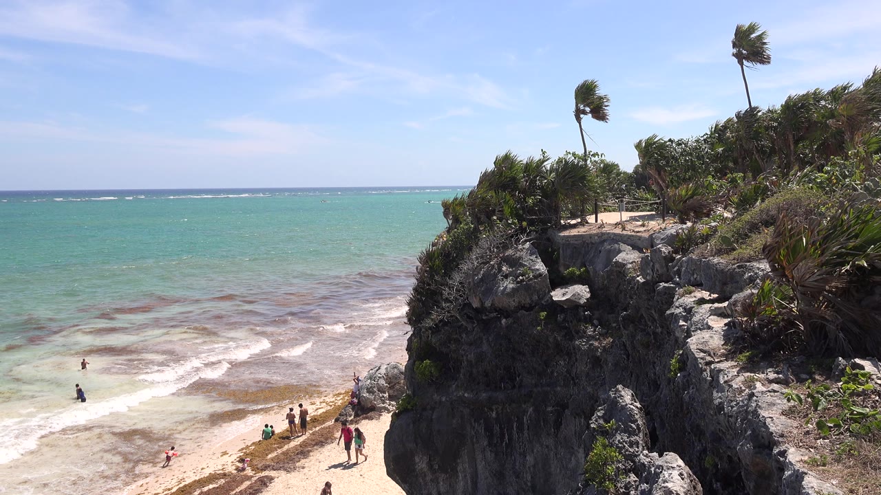 Tulum, Mexico cliffside beach view with the rock face filling the right side, shallow surf spreading across the sand, and tiny people near the waterline.