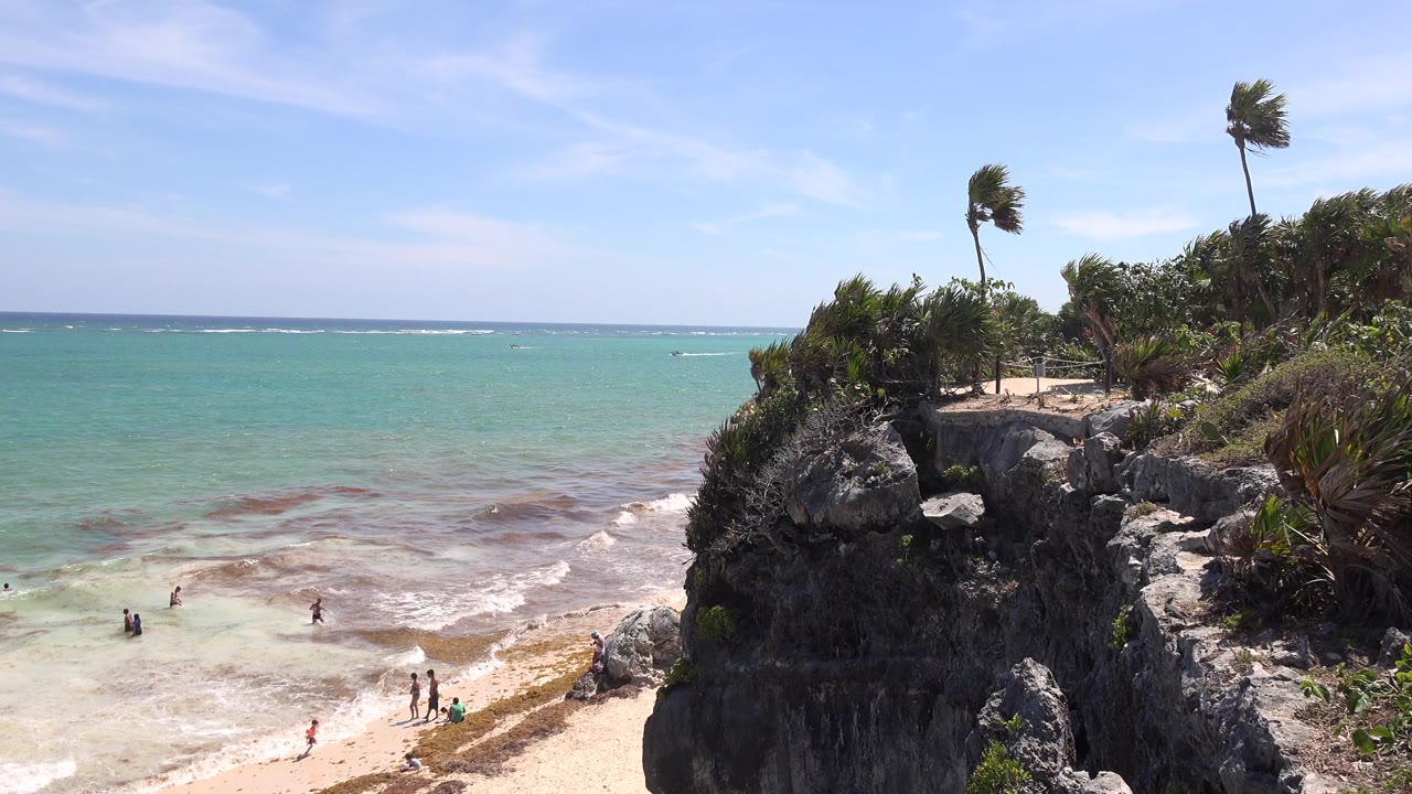 Tulum, Mexico beach view with several small people near the waterline, pale surf spreading over sand, and a rocky vegetated cliff filling the right side of the frame.