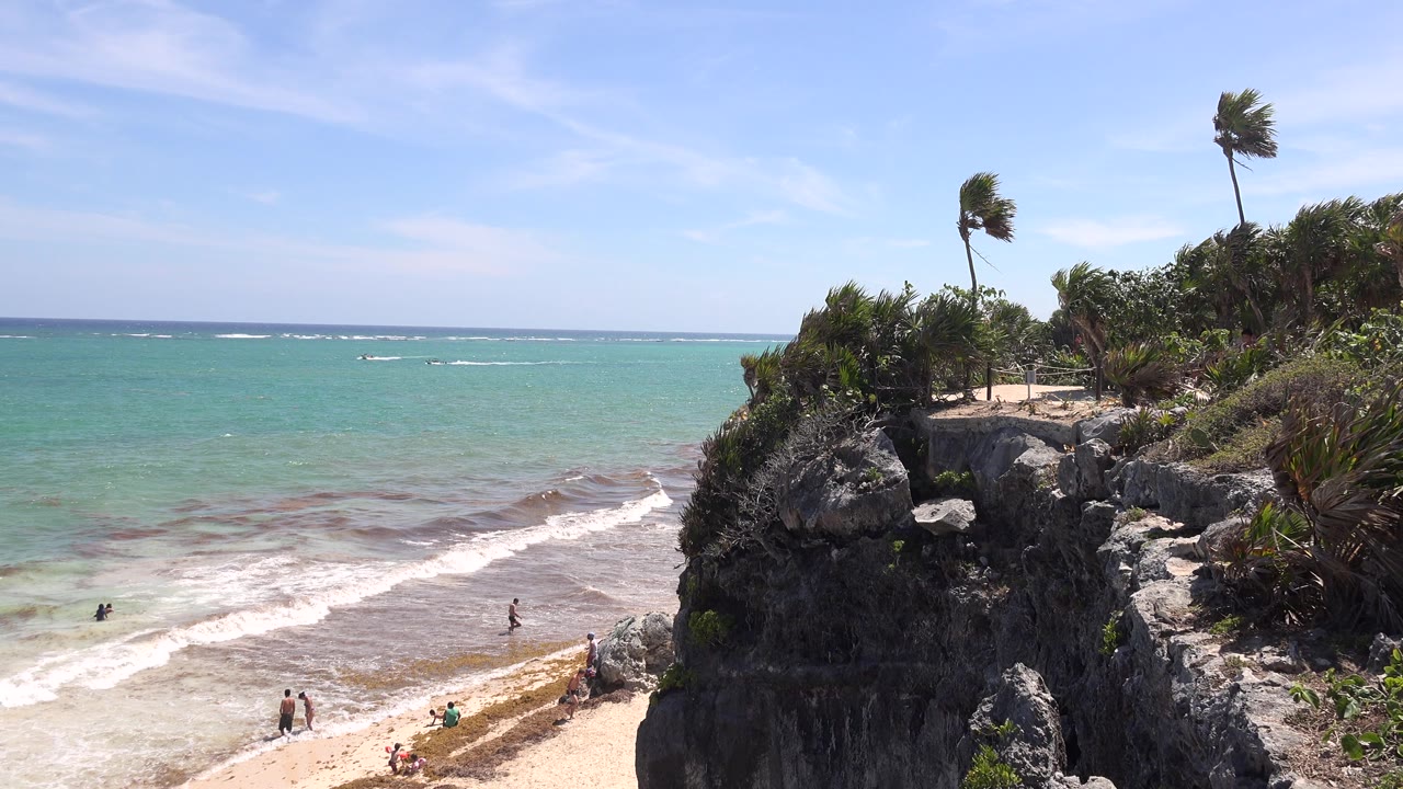 Tulum, Mexico shoreline with white waves reaching the sand, a dark rocky cliff topped by palms, and several small beachgoers visible near the waterline.