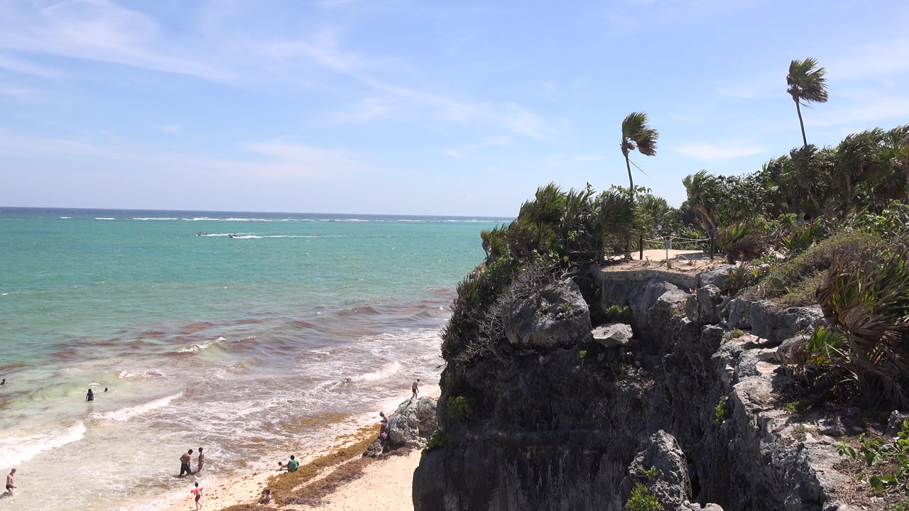 Rocky Tulum shoreline with turquoise water, surf, palms, and beach visitors.