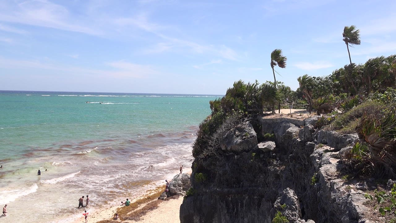 Tulum shoreline view with the rocky cliff prominent on the right, palms leaning in the wind, shallow waves crossing the beach, and a few distant people visible near the waterline.