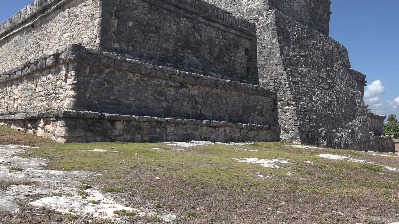 Opening frame shows the lower side of a stone pyramid or ruin in Tulum, Mexico, with rough grass, pale limestone patches, blue sky, and no visible people.