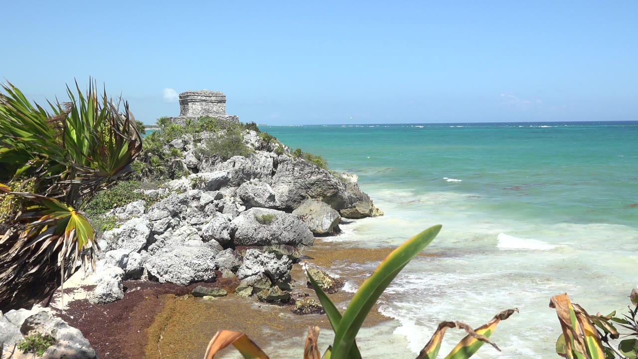 Opening frame of Tulum, Mexico coastline with spiky plants at left, gray rocks below a small stone structure, blue-green water, white surf, and no visible people.