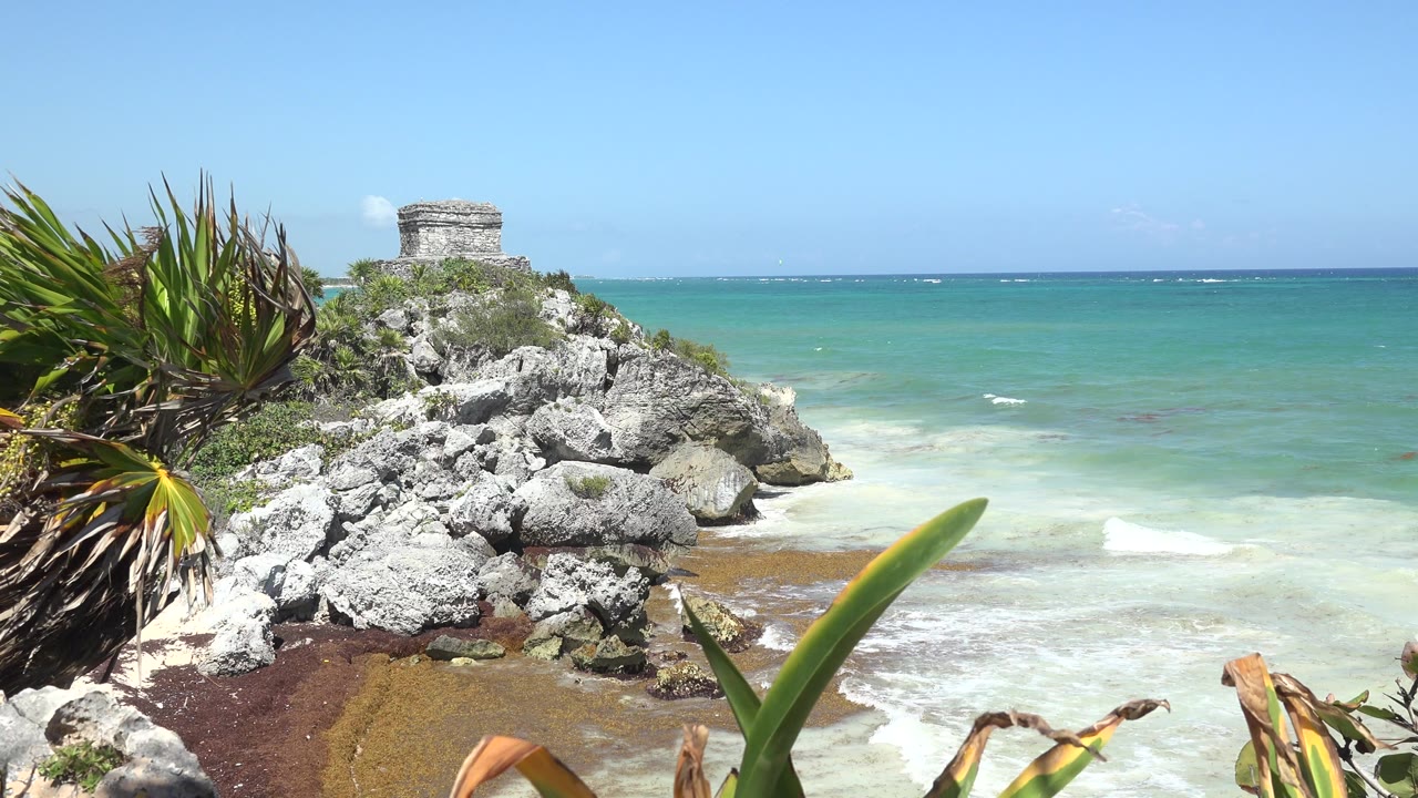 Tulum shoreline view with the stone structure near the upper left, gray rocks across the center, foreground plant leaves, pale surf, blue-green ocean, and no visible people.