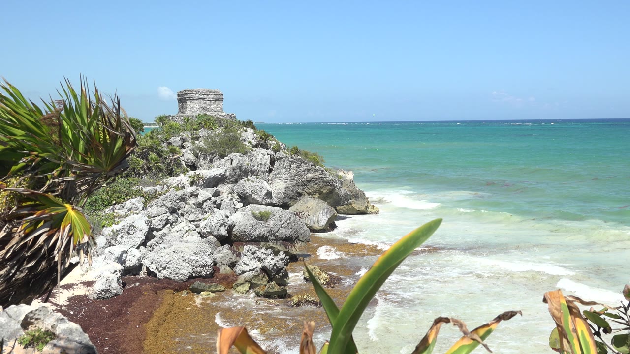 Opening frame of a rocky Tulum, Mexico shoreline with spiky plants at left, pale rocks, a small stone structure above the coast, turquoise water behind it, and no visible people.