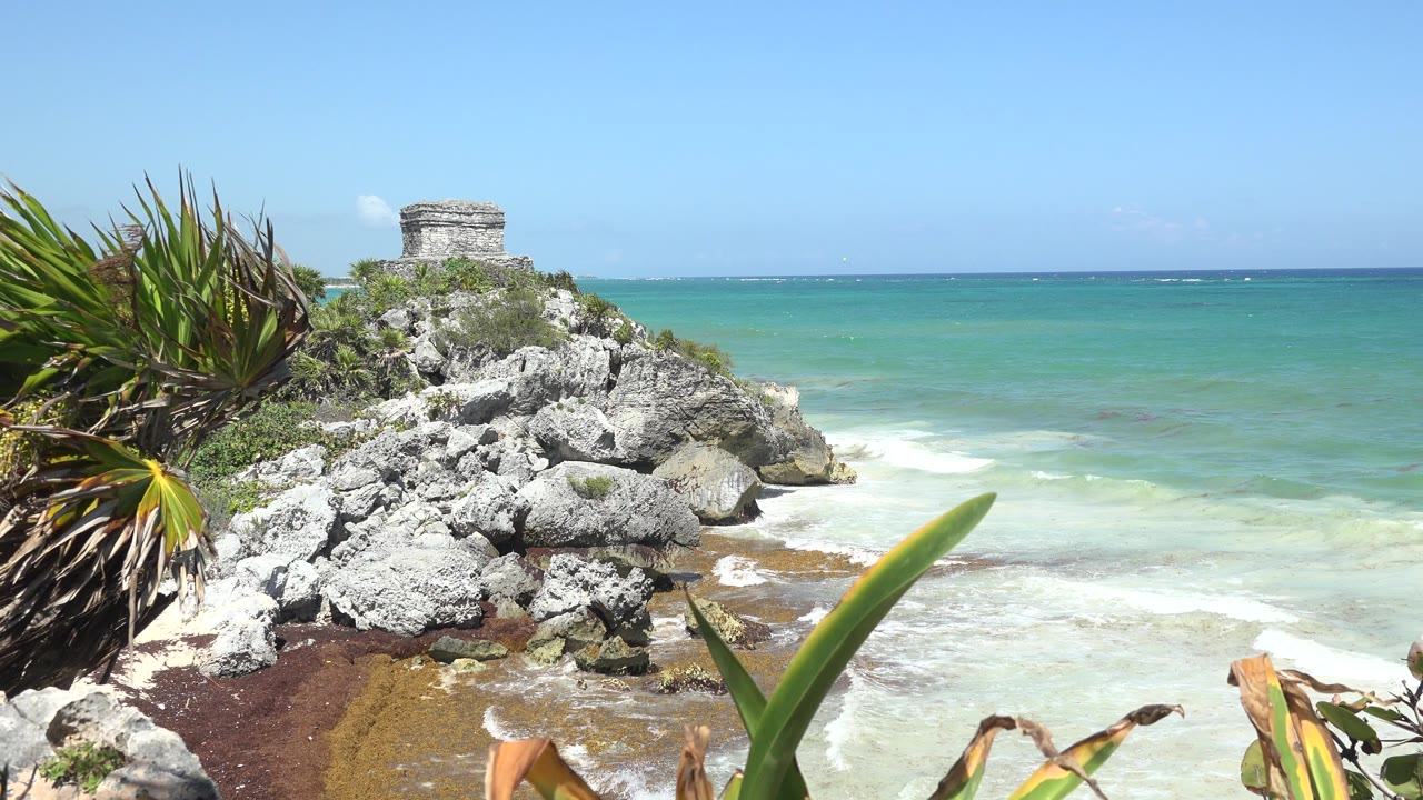 Second frame showing the Tulum, Mexico rocks and small stone structure held near the left side of the frame, with white surf spreading along the shoreline and no visible people.