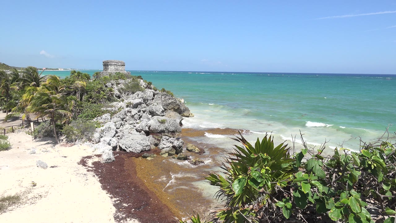 Tulum beach with rocks, palms, blue-green water, and a distant stone structure.