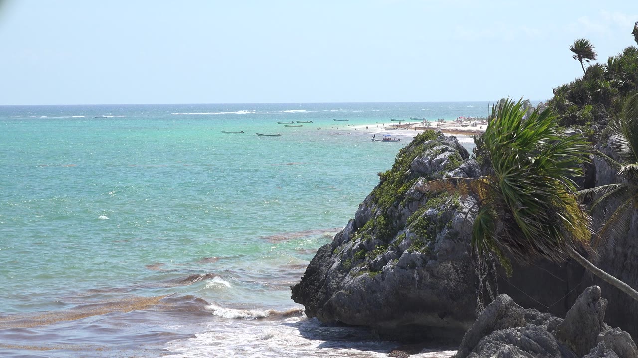 Opening frame of the Tulum, Mexico coast with turquoise water, a dark rocky outcrop at right, green foliage, distant boats, and no visible people.