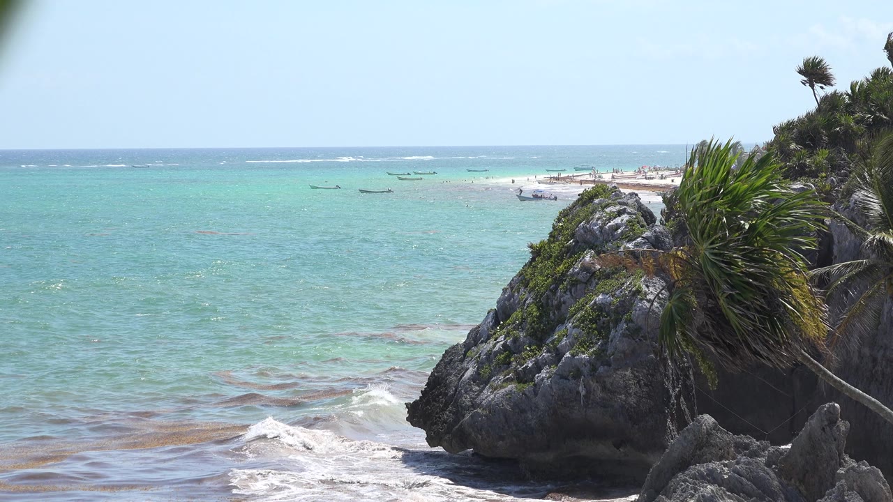 Wave breaks below a rocky point with foliage over bright turquoise water.