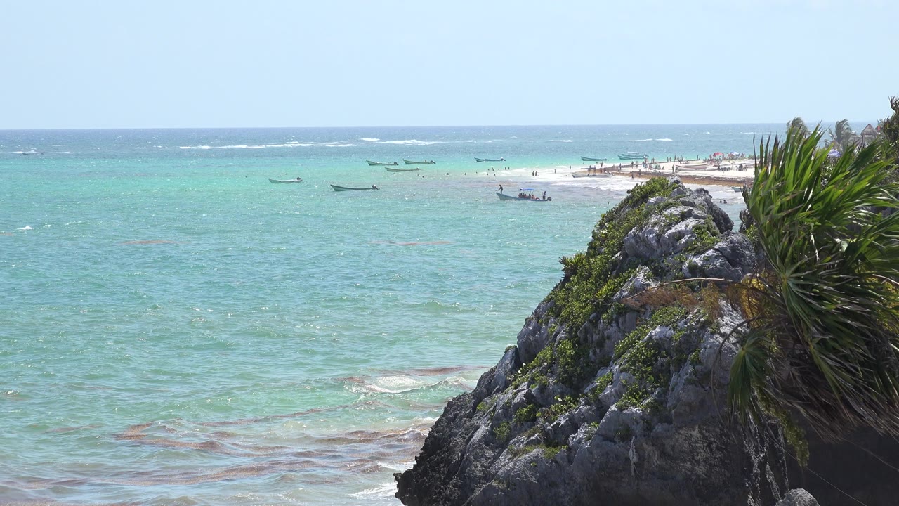 Opening view of the Tulum, Mexico shoreline with turquoise water, small anchored boats near a pale sandbar, dark rocks and green plants at right, and no visible people.