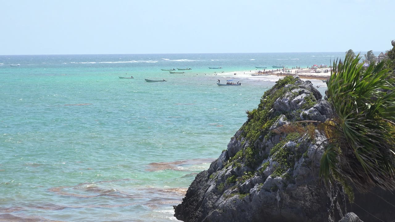 Blue-green water, distant boats, sandy strip, and right-side coastal rock.
