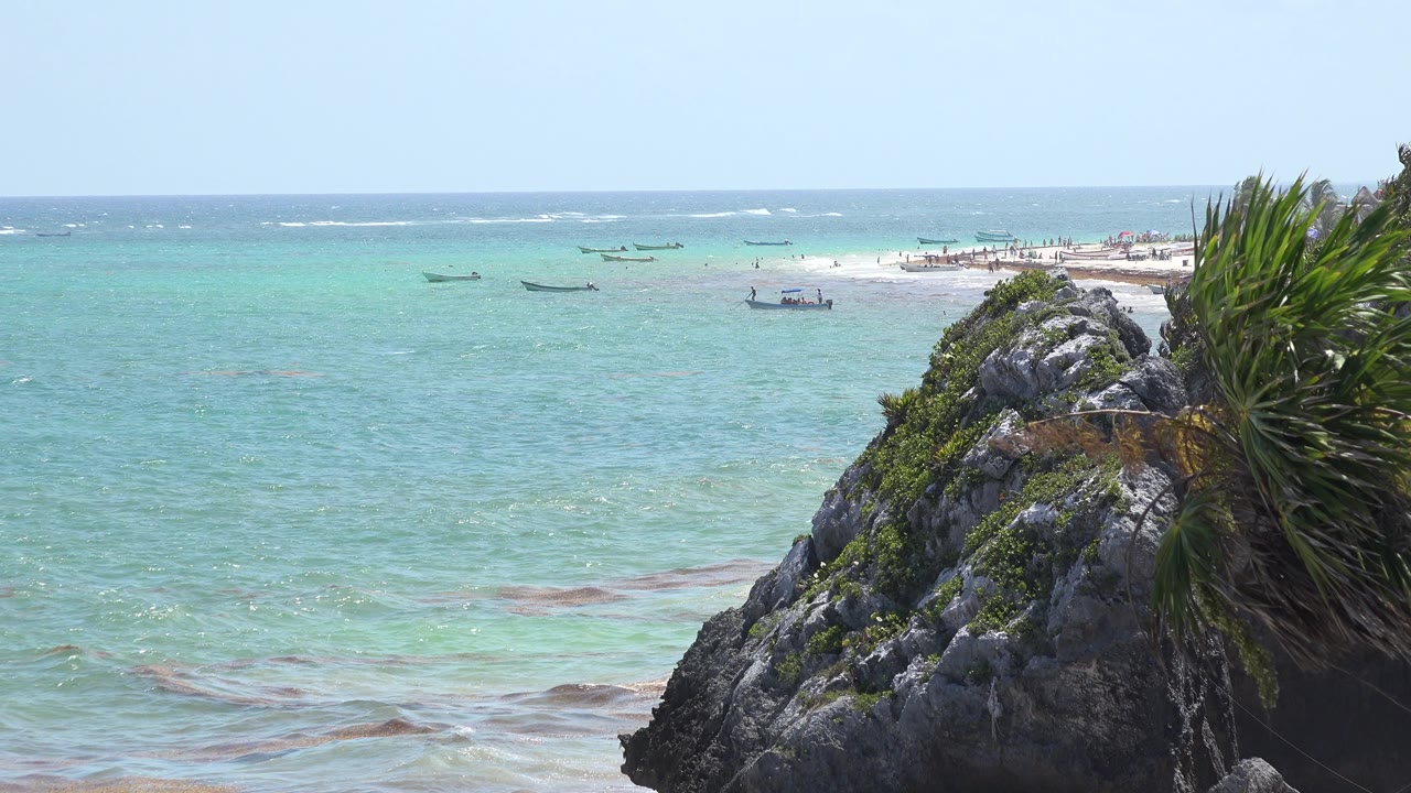 Opening frame of Tulum, Mexico coastline with a dark rocky outcrop and green plants at right, turquoise water across the middle, distant beach, small boats offshore, and no visible people.