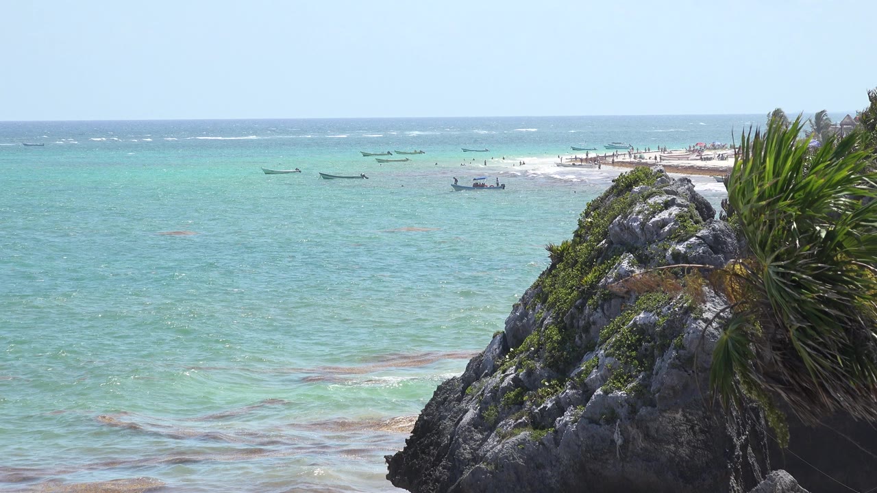 Opening frame of a Tulum, Mexico coastal view with turquoise water, a dark rocky foreground, green palm fronds at right, small boats offshore, and no visible people.