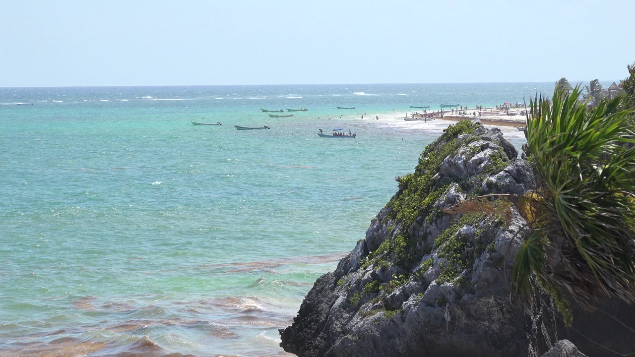 Opening view of the Tulum, Mexico coast with turquoise water, a rocky plant-covered foreground, small boats near the sandbar, and no visible people.