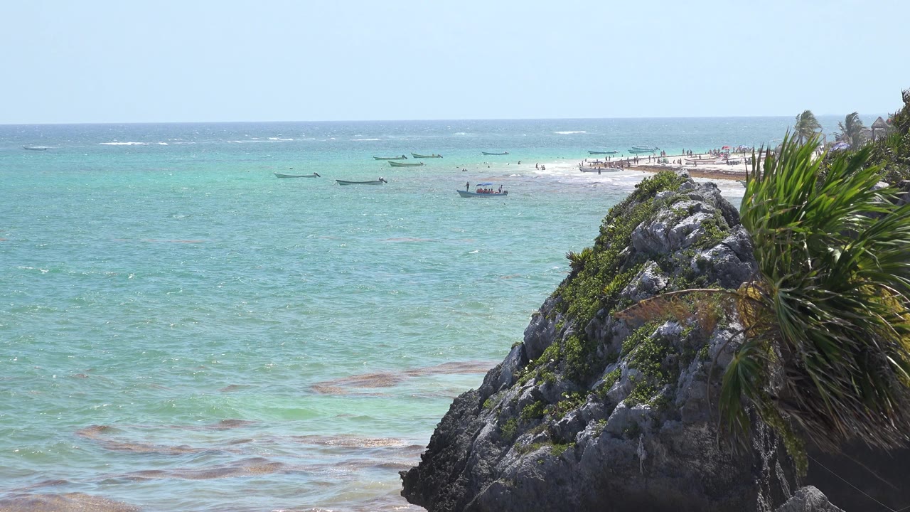 Rocky coastal foreground beside pale turquoise water and distant boats