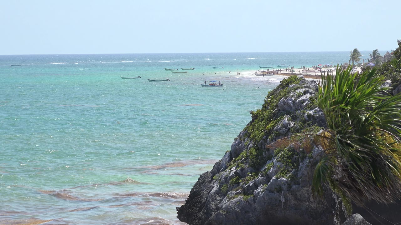 Tulum, Mexico beach view with sunlit turquoise water, a foreground rock and plant cluster at right, a pale sand area beyond, small boats offshore, and no visible people.