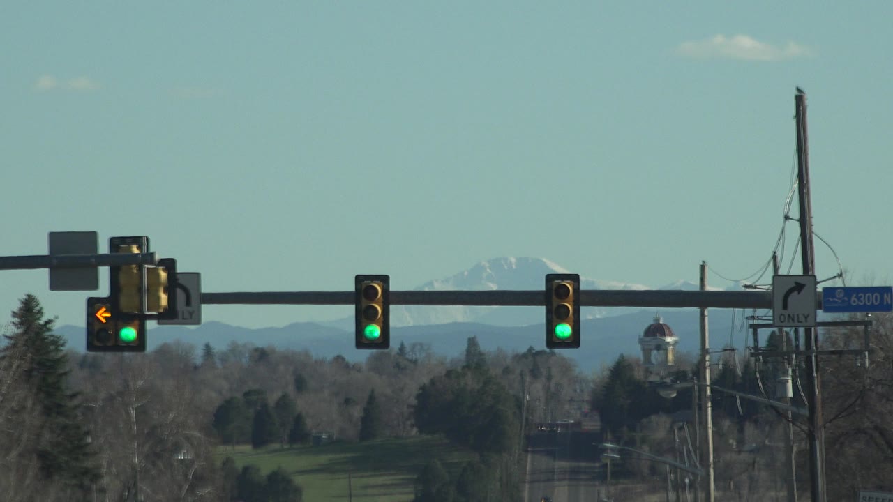Opening frame of Denver traffic signals showing green lights, a yellow left-turn arrow, blue street signage, utility poles, bare trees, and distant snow-capped mountains with no visible people.