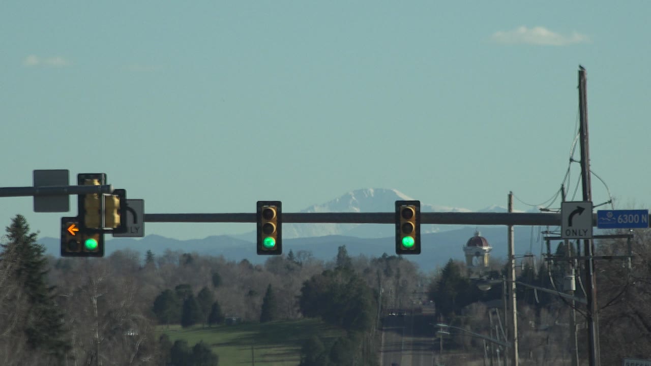 Later Denver frame with two green traffic lights centered along the signal arm, the left-turn arrow still lit at left, roadside poles, winter-bare trees, and snowy mountains in the distance, with no visible people.