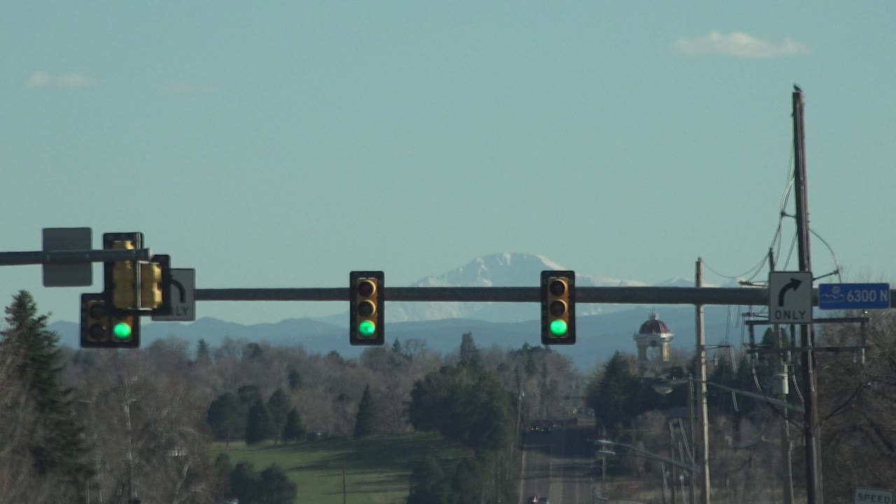 Mid-clip Denver view showing the overhead signal arm crossing the frame, green lights glowing in the foreground, a small tower-like structure near the road, utility lines, trees, and distant mountains, with no visible people.