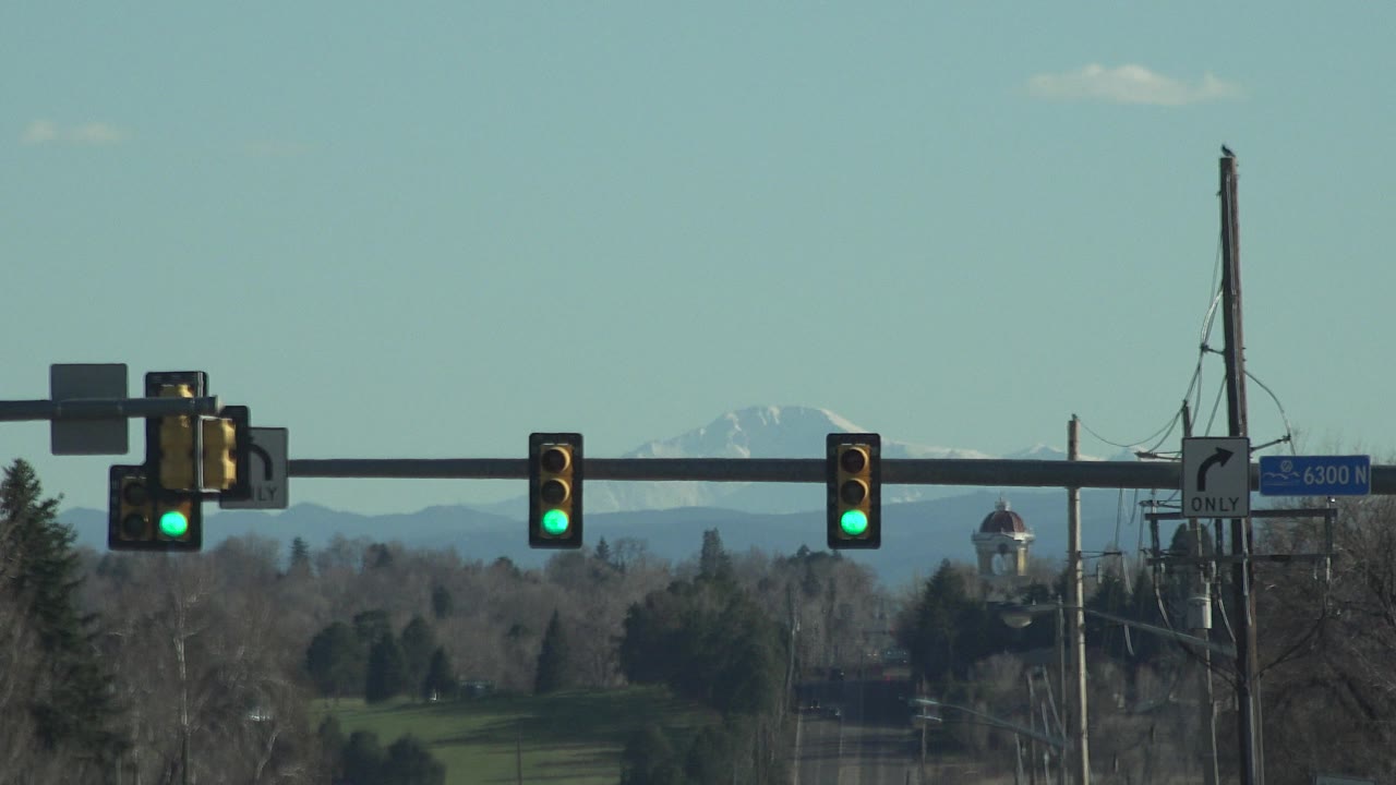A traffic signal arm with green lights and roadside signs frames a long Denver street view, with trees and a hazy distant horizon and no visible people.