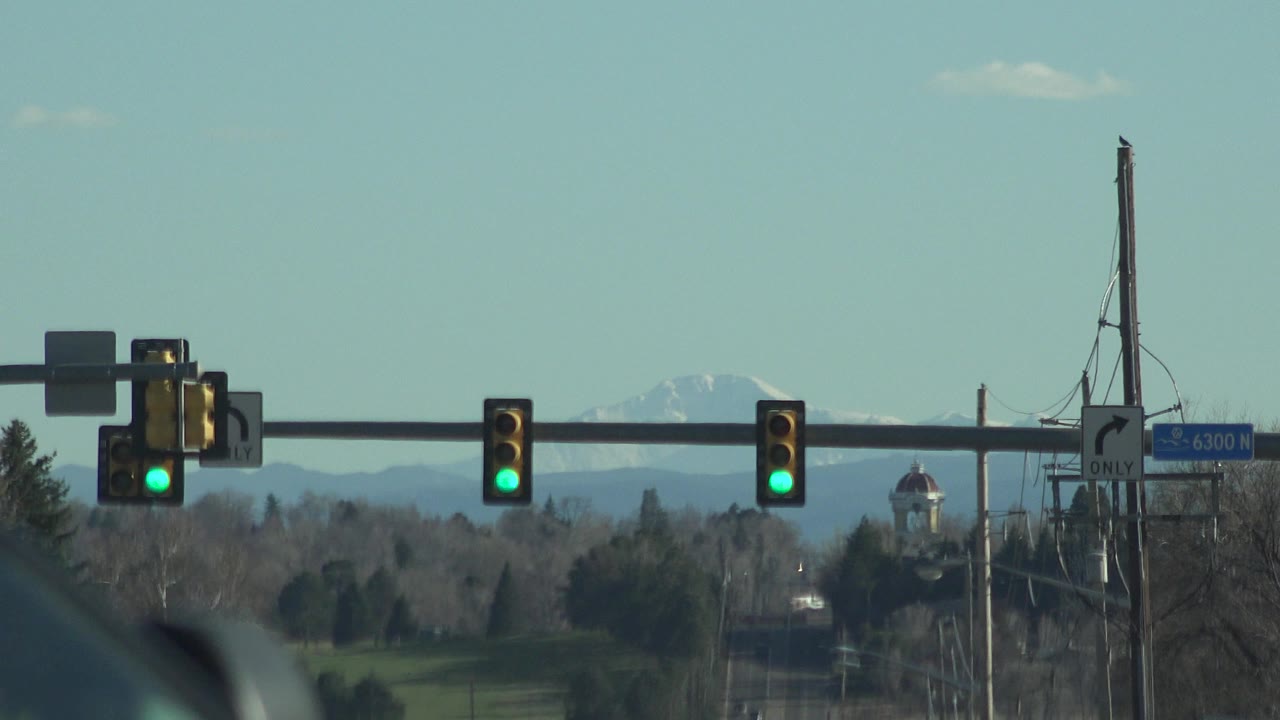 Green traffic lights span a Denver road view with trees, utility poles, a pale sky, distant snow-capped mountains, and no visible people at 0:00.
