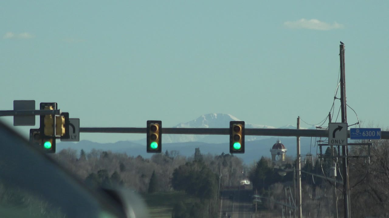A slightly shifted Denver road view shows green signal heads, roadside utility poles, trees, a small distant tower or dome, mountains, and no visible people at 0:01.