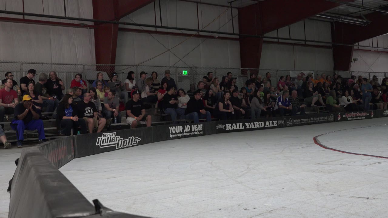 Indoor roller derby rink with spectators on bleachers behind rink boards.