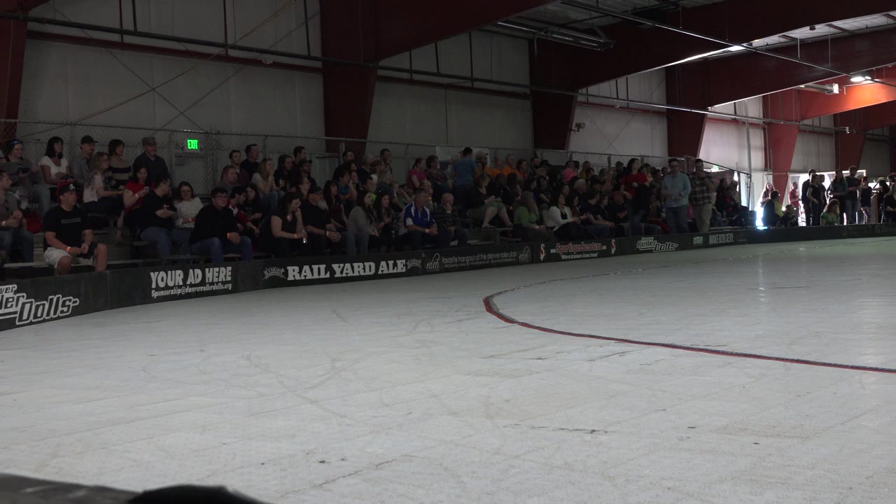 Crowd seated behind rink boards beside an empty indoor rink.