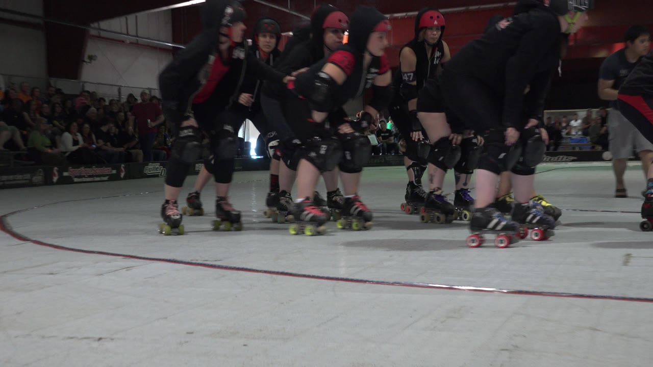 At 0:01 in Denver, Colorado, a group of black-clad roller skaters remains close to the low camera while a person in shorts passes in the background near the spectators.