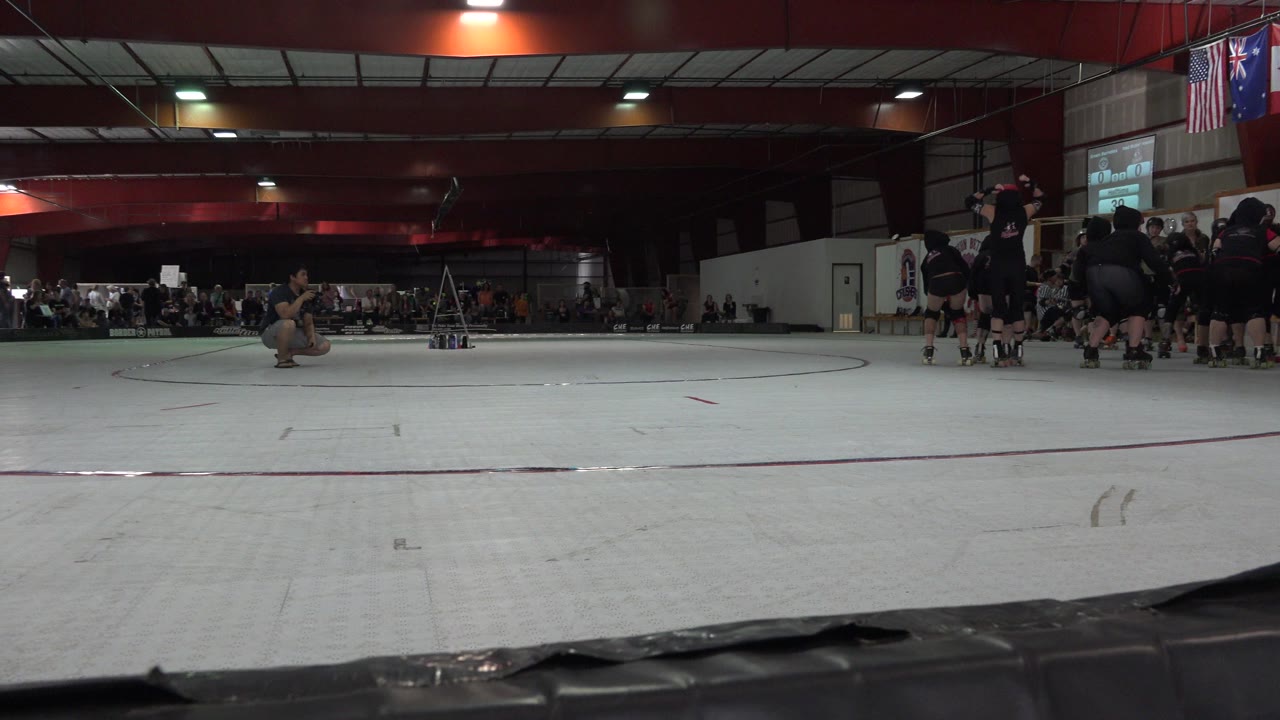 Denver roller derby warmup frame with skaters in dark gear grouped near the right wall, open rink floor in front, and seated spectators along the far side.