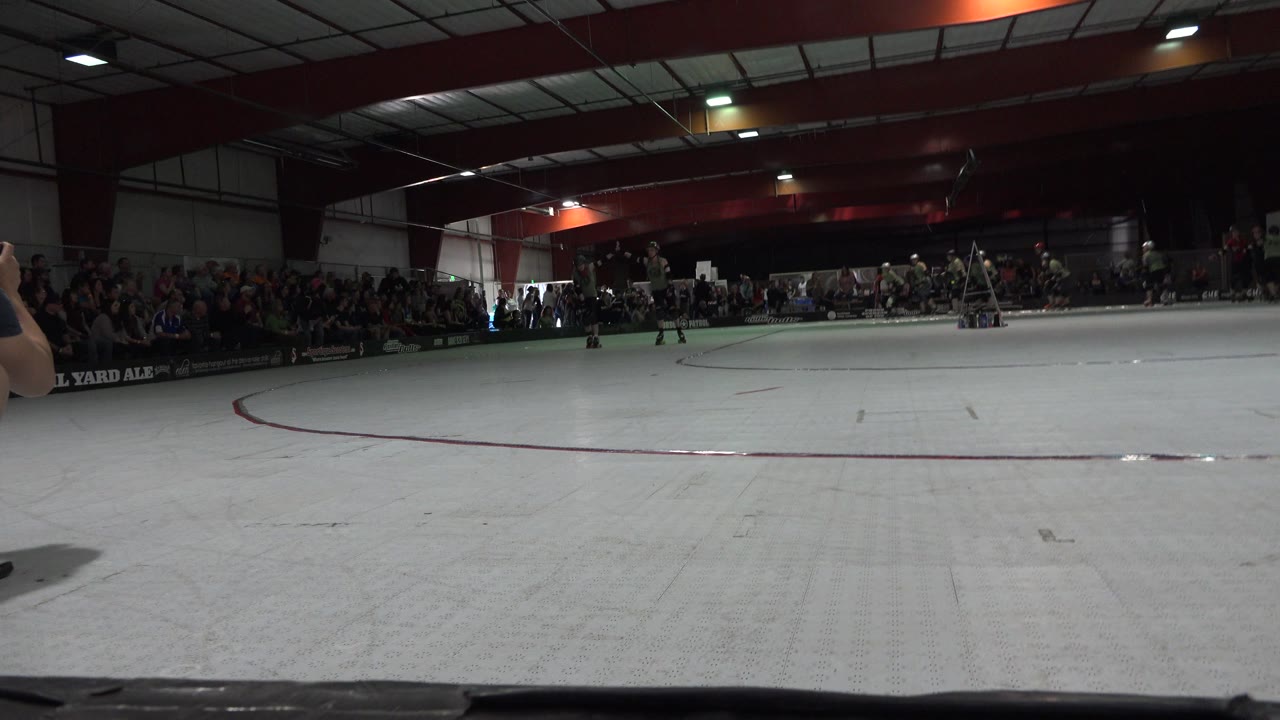 Low rinkside view in Denver with the pale skating surface across the foreground, spectators in the background, and a raised arm at the right edge.