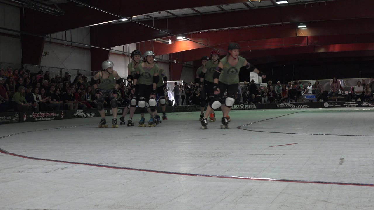 A group of women in green and black roller derby gear stand and begin skating on an indoor track in Denver, with spectators seated behind the rink boards.