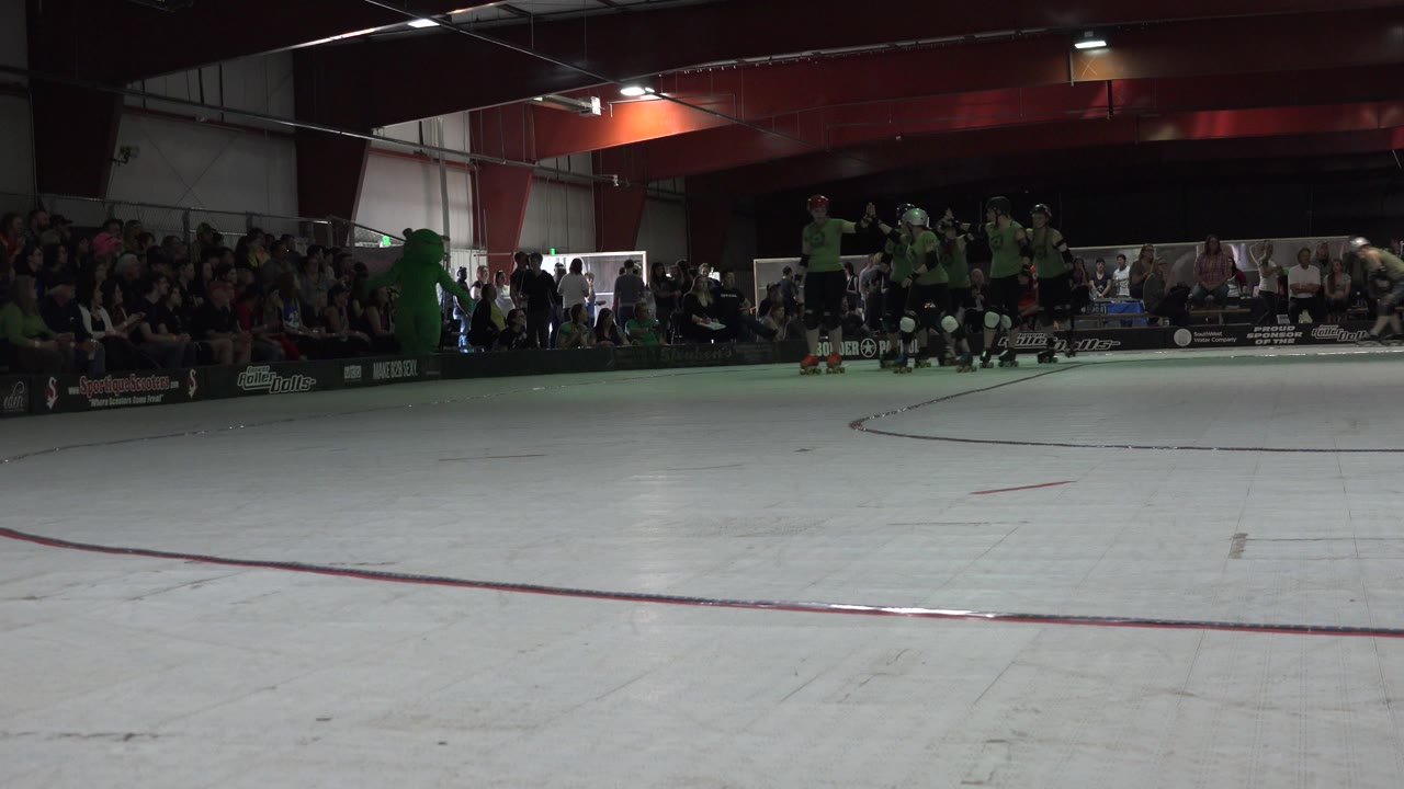 A cluster of helmeted skaters in green shirts gathers along the edge of the Denver indoor court while a crowd sits behind the boards.