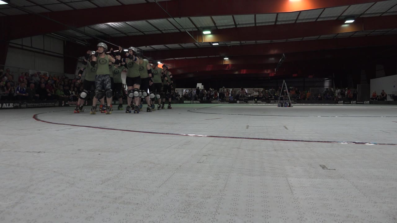 Helmeted skaters in olive shirts remain grouped near the left side of the Denver indoor rink while a seated crowd fills the dim background.