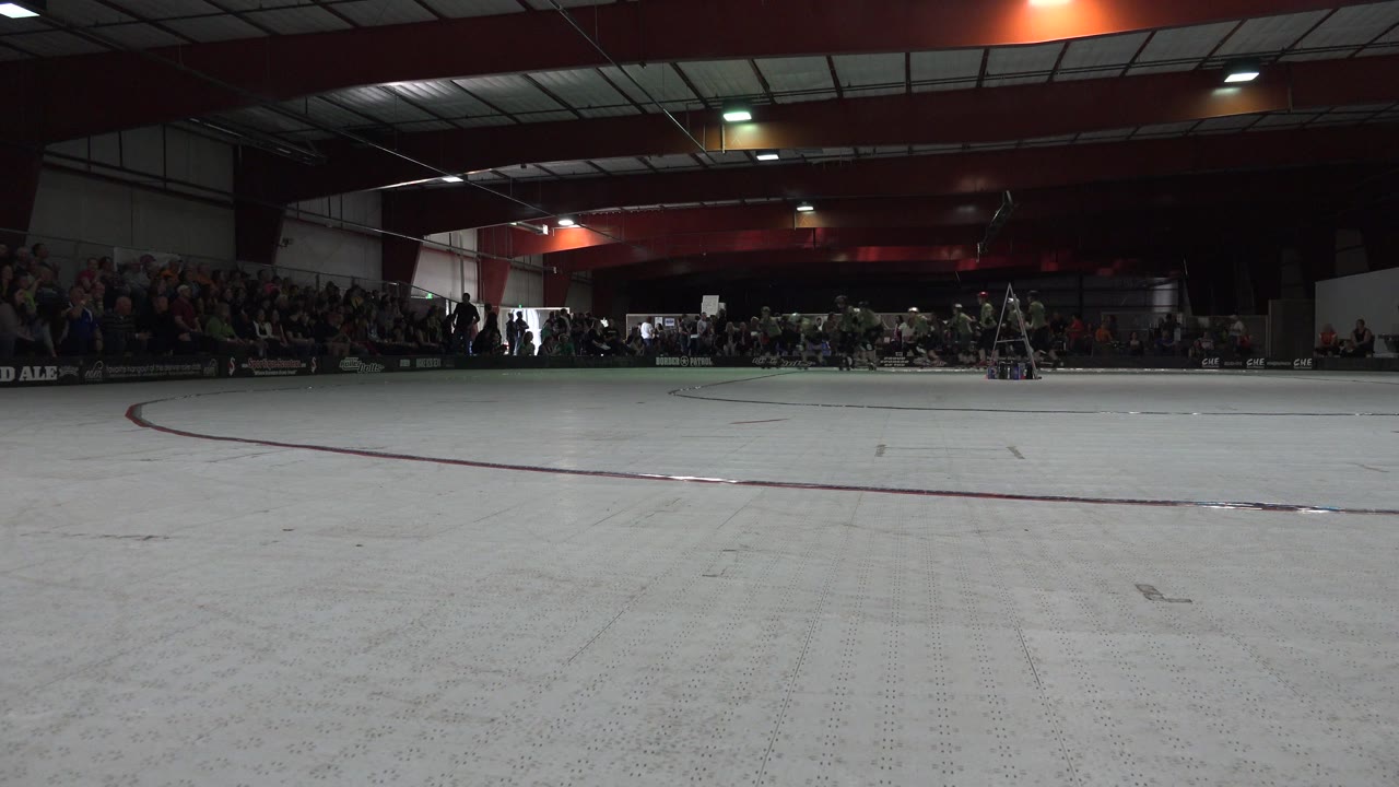 Wide indoor roller rink with spectators seated along the far wall.