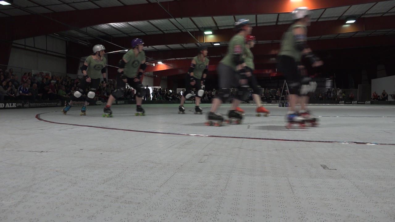 Helmeted roller skaters pass close to a low camera inside an arena.