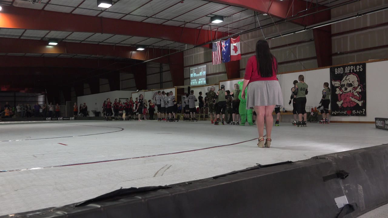 Woman stands rinkside facing athletes during an indoor sports event.