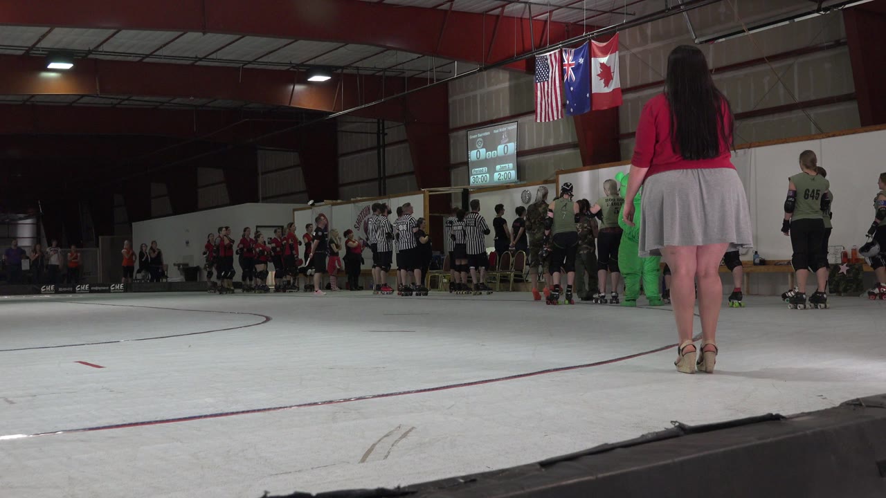 Low ground-level view in Denver, Colorado, with a woman in a red top and white skirt facing skaters, officials, spectators, and national flags inside an indoor rink.