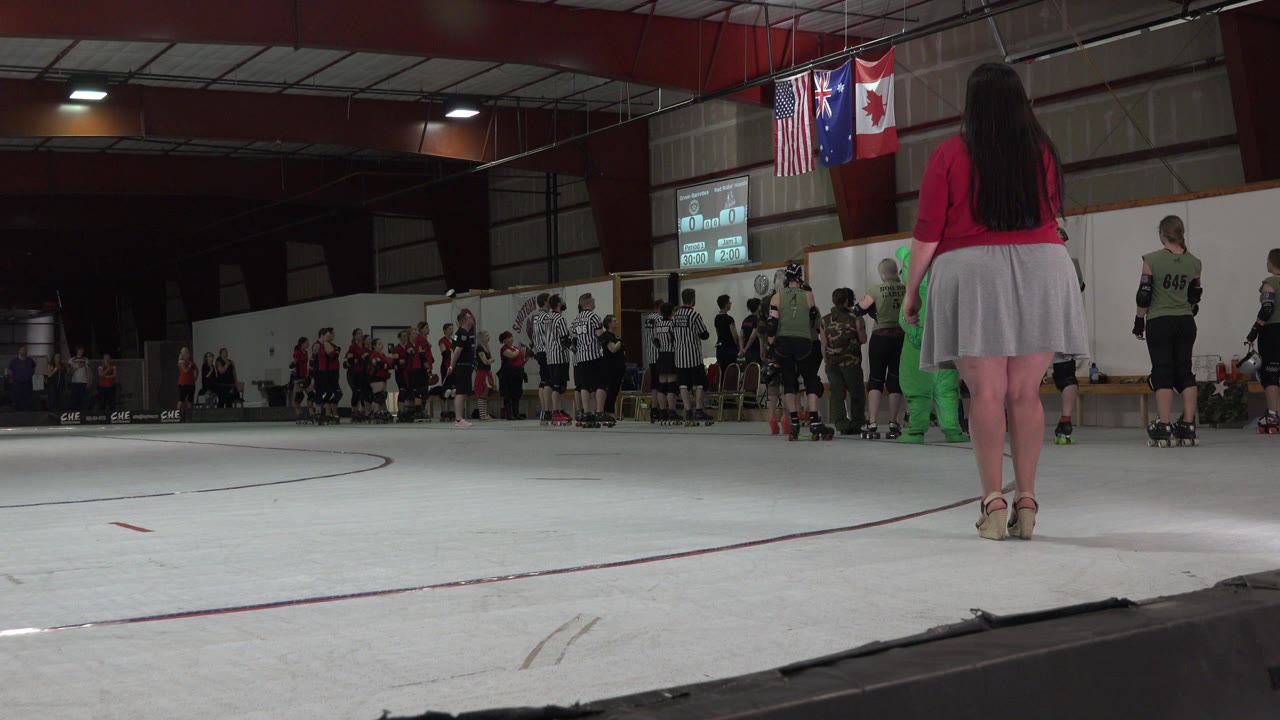 Woman in red top faces a crowd on an indoor rink beneath hanging flags.