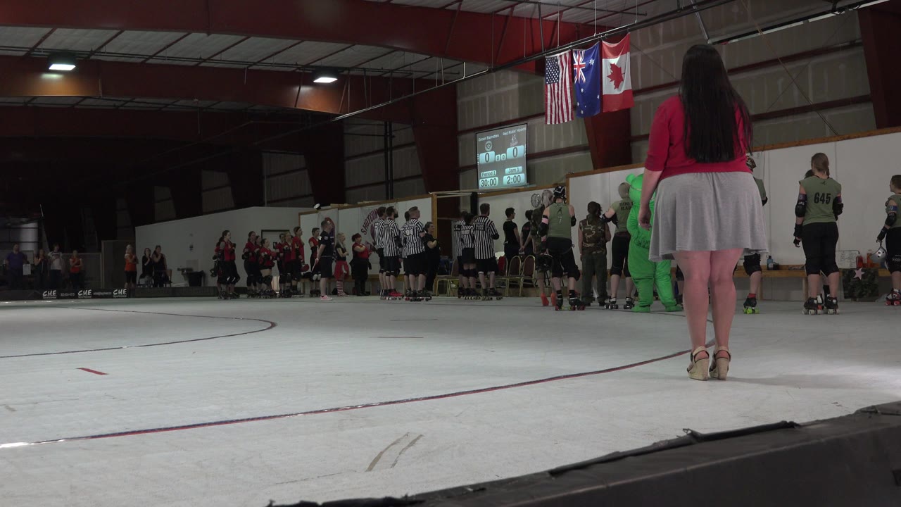Low court-level frame in Denver shows a woman in a red top and gray skirt facing players and referees gathered near the far wall, with spectators and hanging flags in the background.