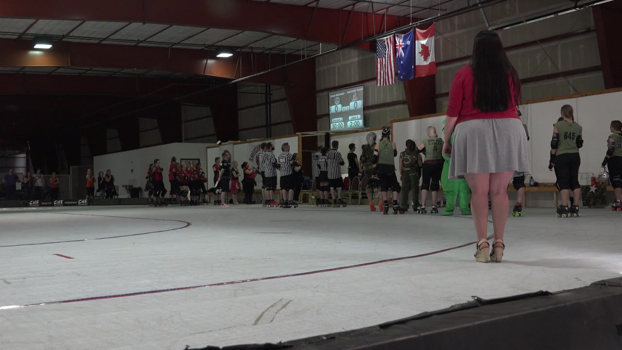 At 0:00 in Denver, a low view from beside the rink shows athletes, referees, spectators, and American and Canadian flags hanging above the boards.