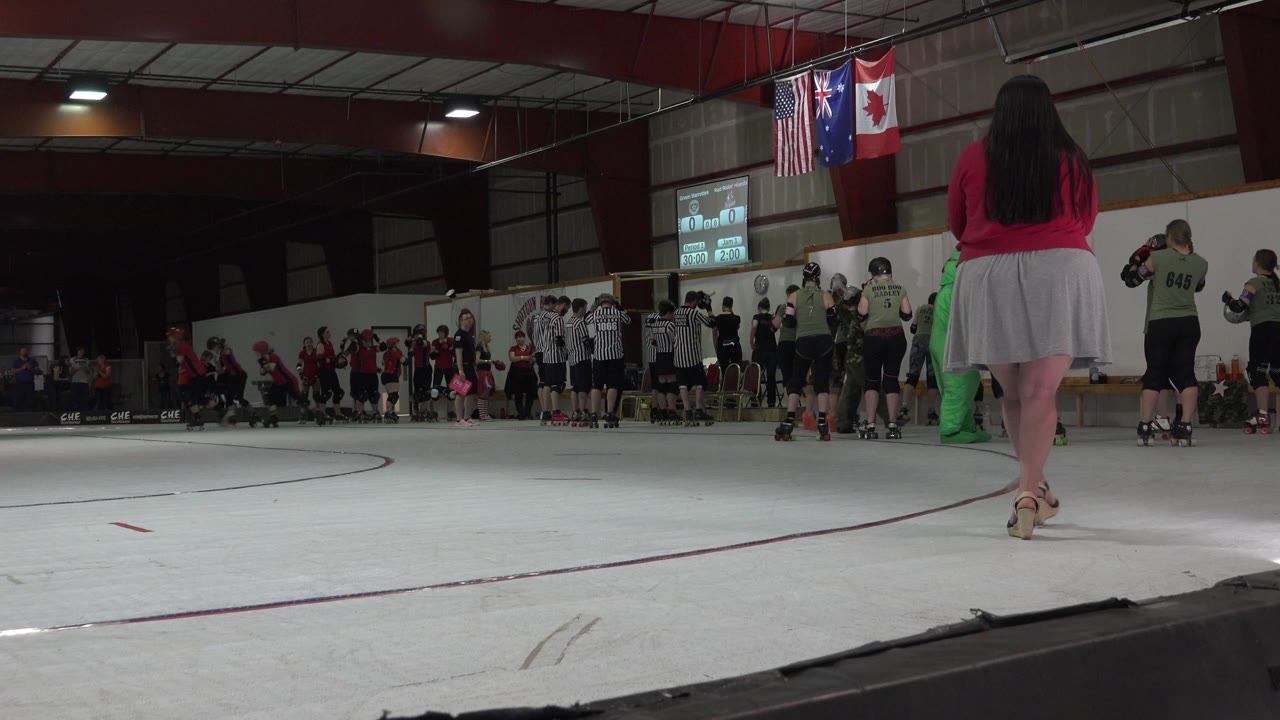 Roller skaters and officials gather on an indoor rink as a woman crosses the foreground.
