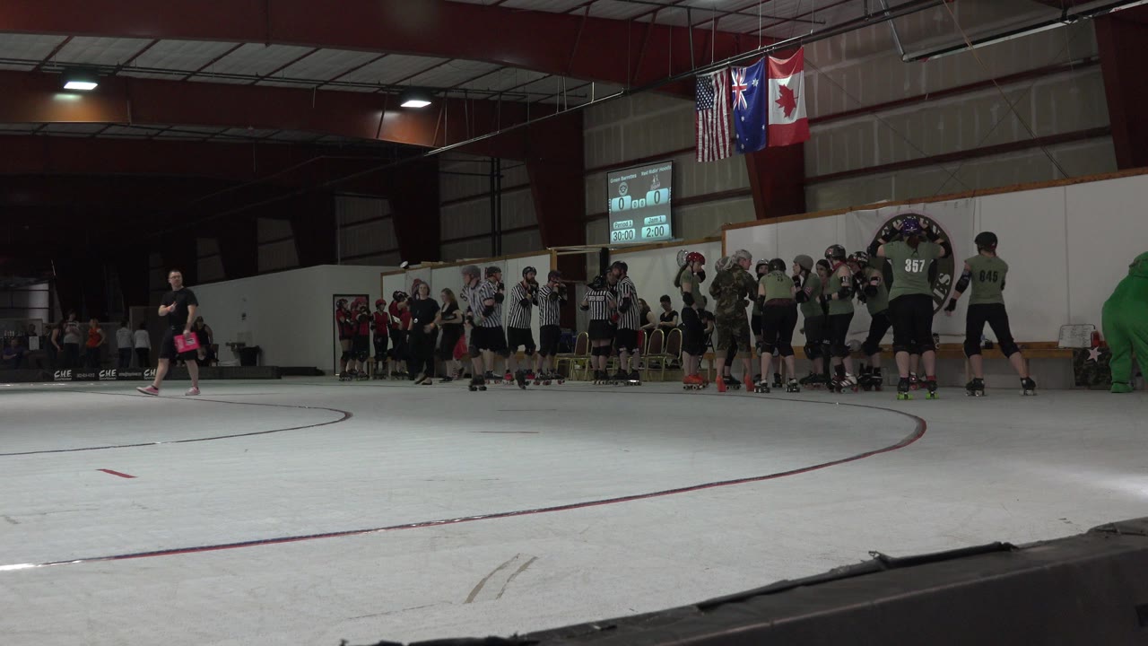 Denver indoor skating game frame showing a wide rink foreground, clustered helmeted players, striped referees, flags overhead, and spectators along the back wall.